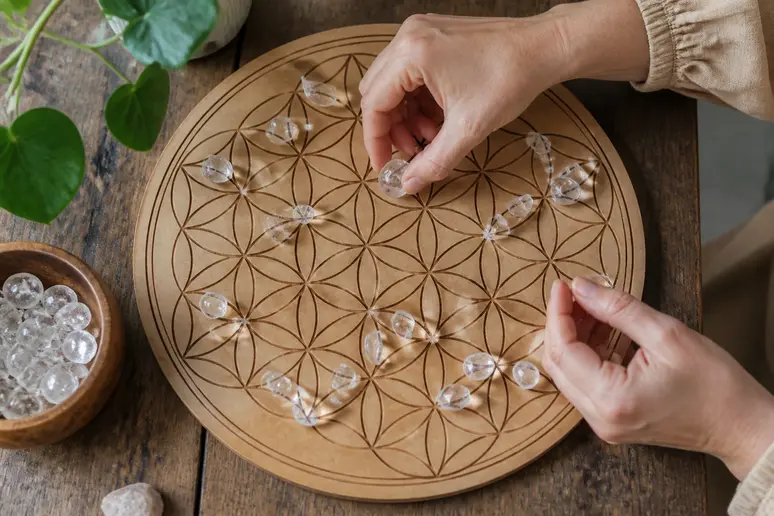 Des mains déposent délicatement des cristaux polis sur une grille en bois gravée représentant la fleur de vie.