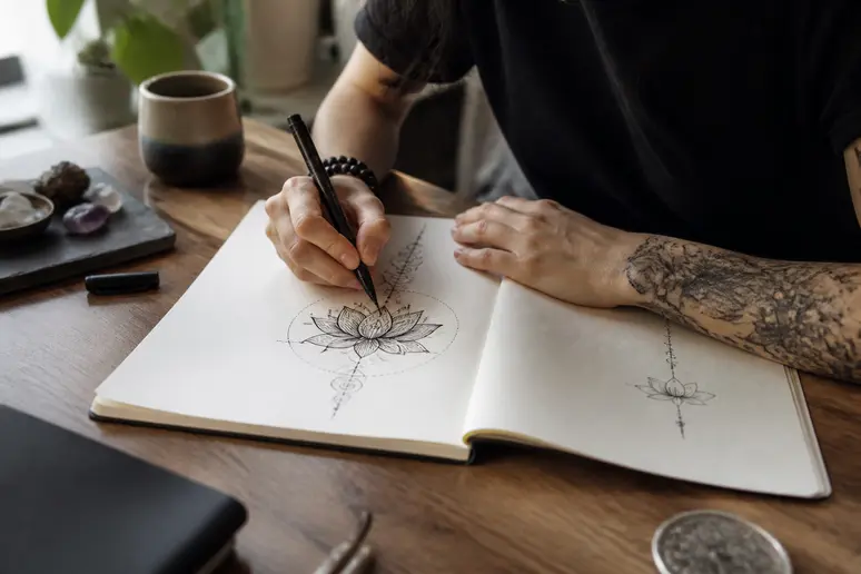 Personne dessinant un motif de tatouage karma et un symbole spirituel dans un carnet.